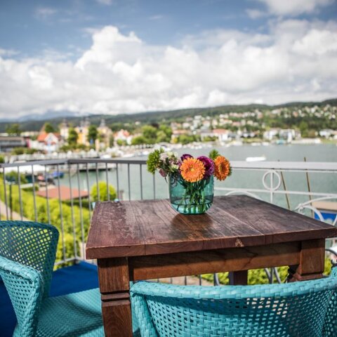 <p>Balkon mit rustikalem Holztisch mit Blumen und türkisen Stühlen in der Bucht von Velden am Wörthersee.</p>